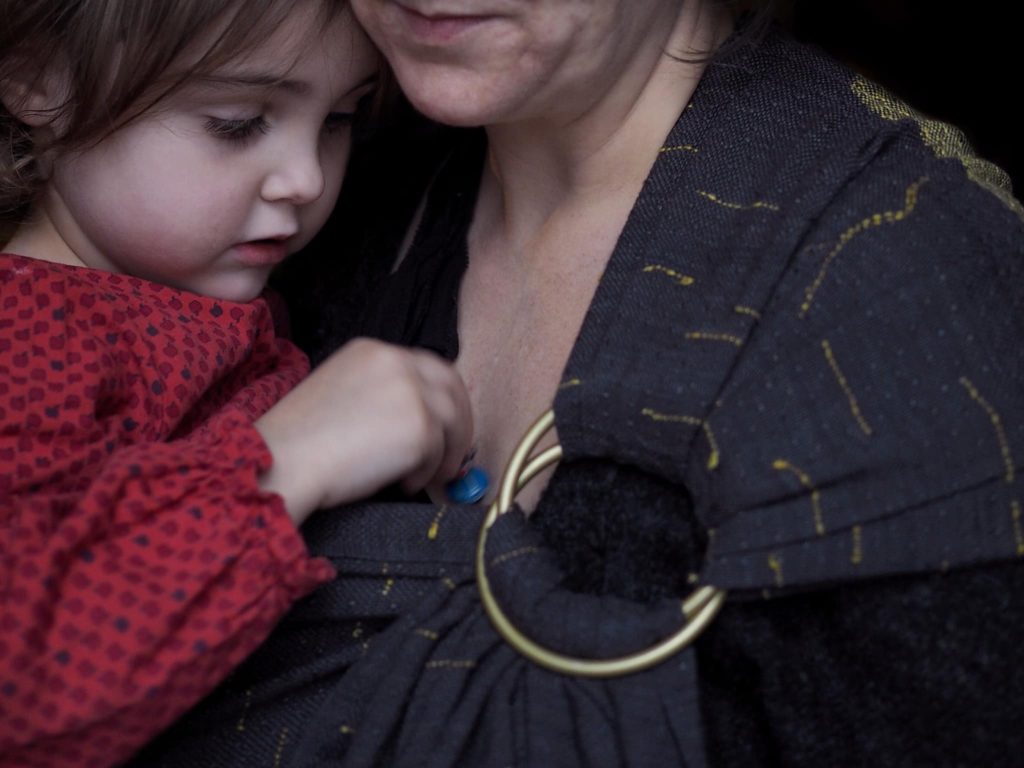 a white woman carries her preschooler child in a black and gold textured ring sling