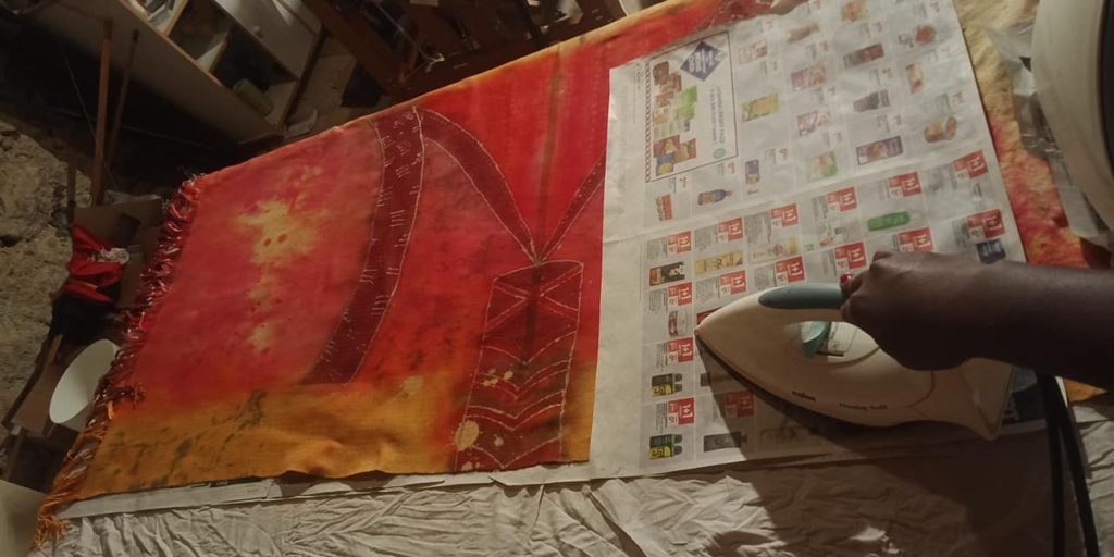 Image of black hand, ironing a wrap in between newspaper sheets. wrap on a table, wmdyed in gold, reds, brown. Maroon and gold fringes with white 'beads'.